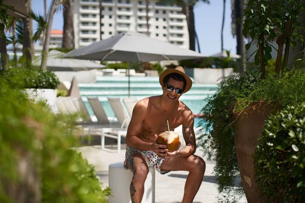 Shirtless man in a fedora and sunglasses drinking out of a whole coconut
