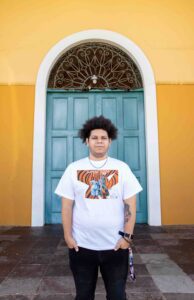 art student jehiel franco torres wearing shirt designed by him in front of yellow wall and blue door